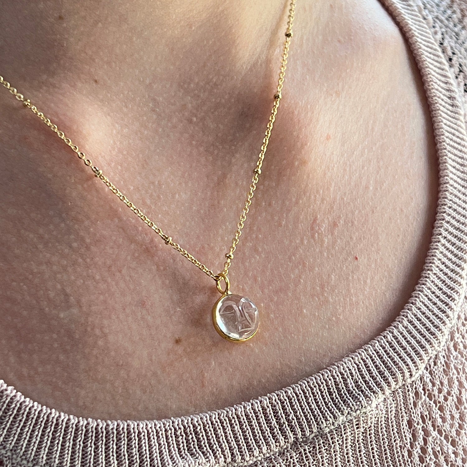 A gold-plated satellite chain necklace with a carved rock crystal moon face pendant wornb around the nexk or a perosn with a beige top.