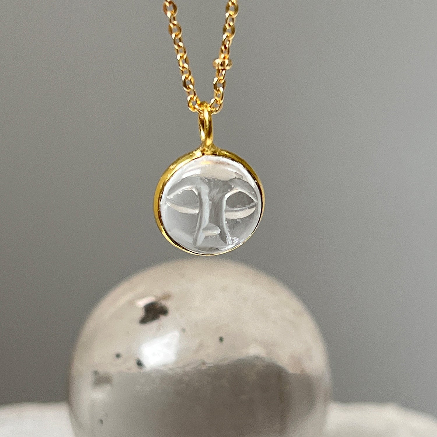 A gold-plated satellite chain necklace with a carved rock crystal moon face pendant against the background of a stone.