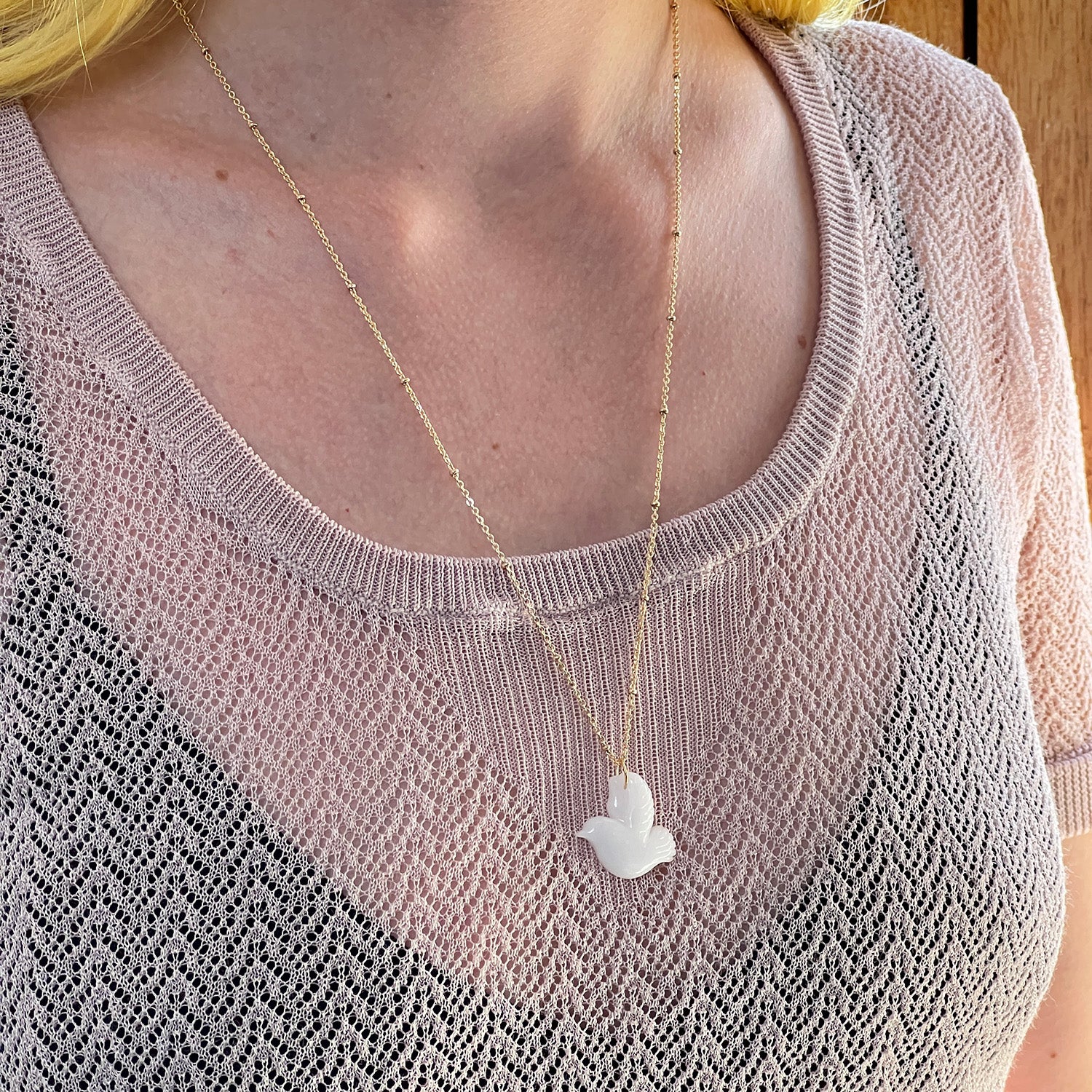 A closeup of a model wearing white dove necklace made from agate, with a gold chain. The model is wearing a light knitted top.