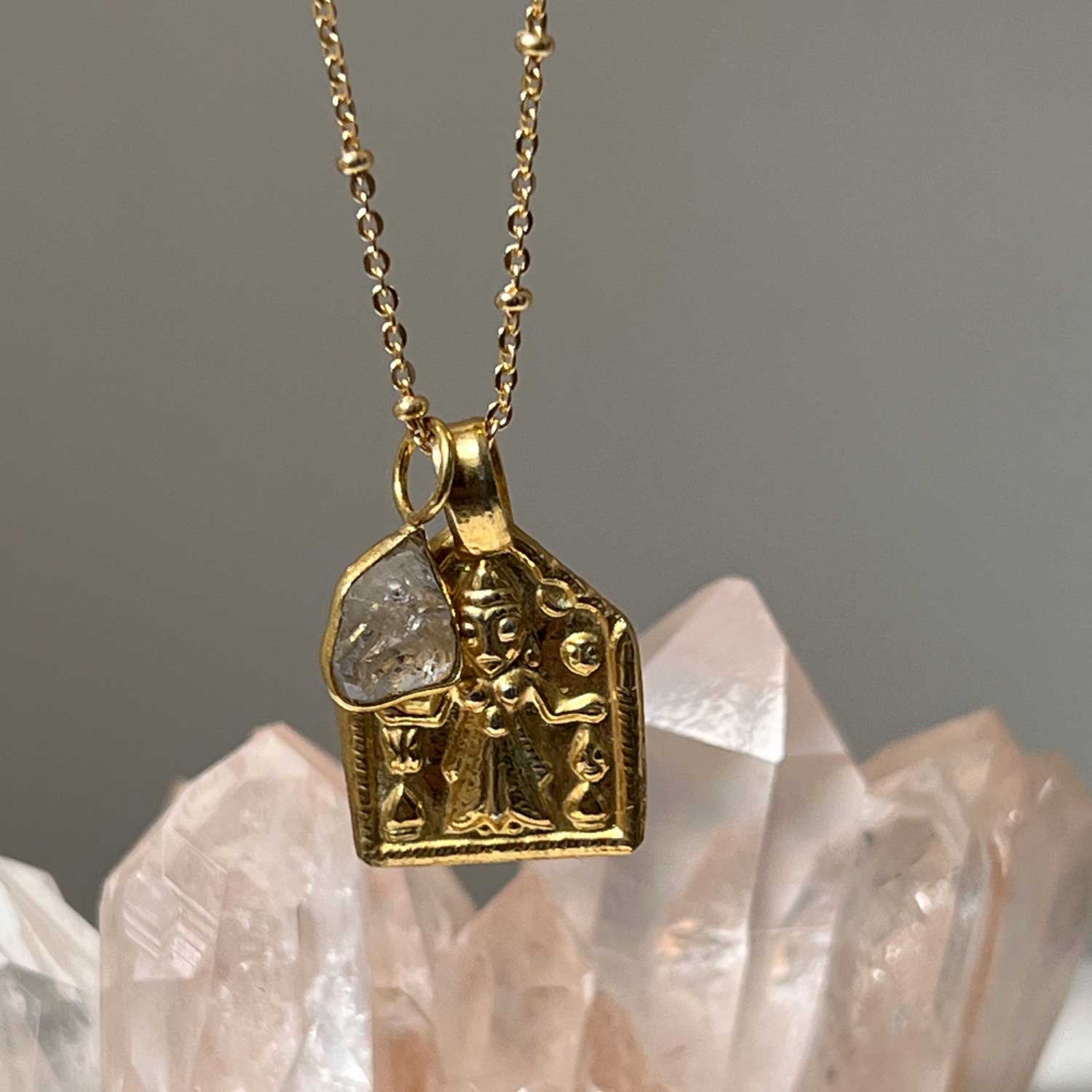 A gold-plated pendant featuring Goddess Lakshmi with a Herkimer Diamond Slicewith pink crystals in the background