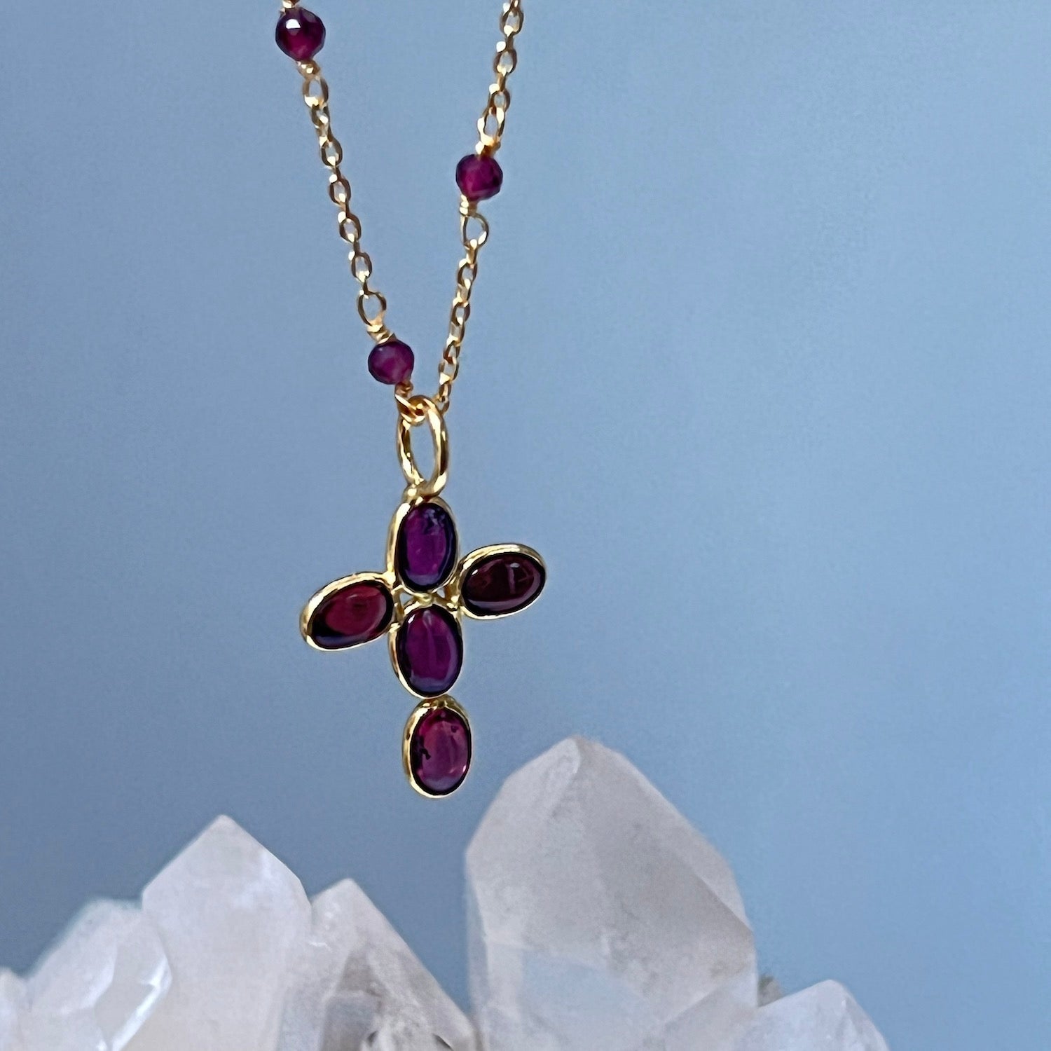 Gold necklace with a cross pendant featuring red garnet gemstones displayed next to a crystal