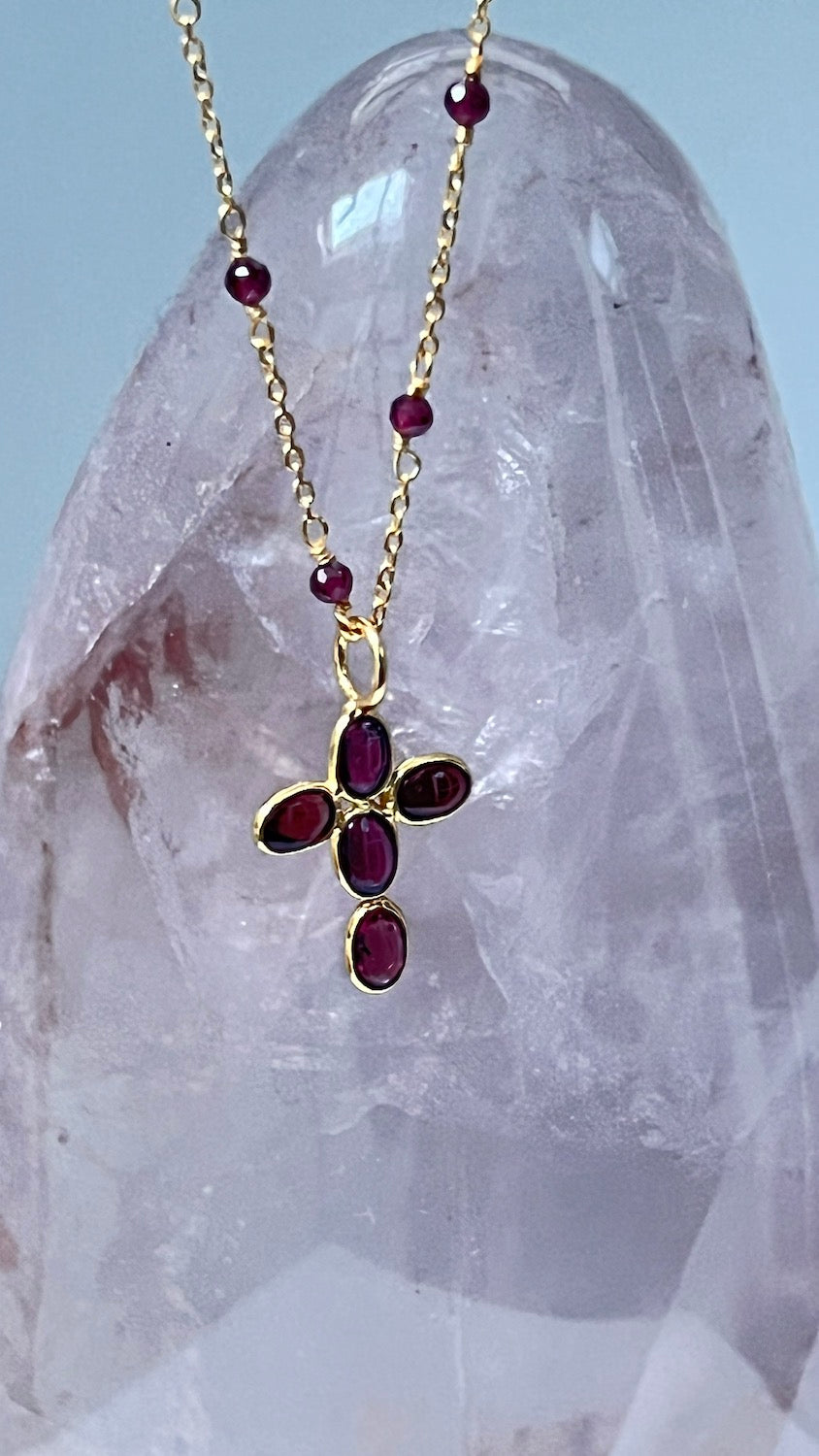 Gold necklace with a cross pendant featuring red garnet gemstones displayed next to a crystal