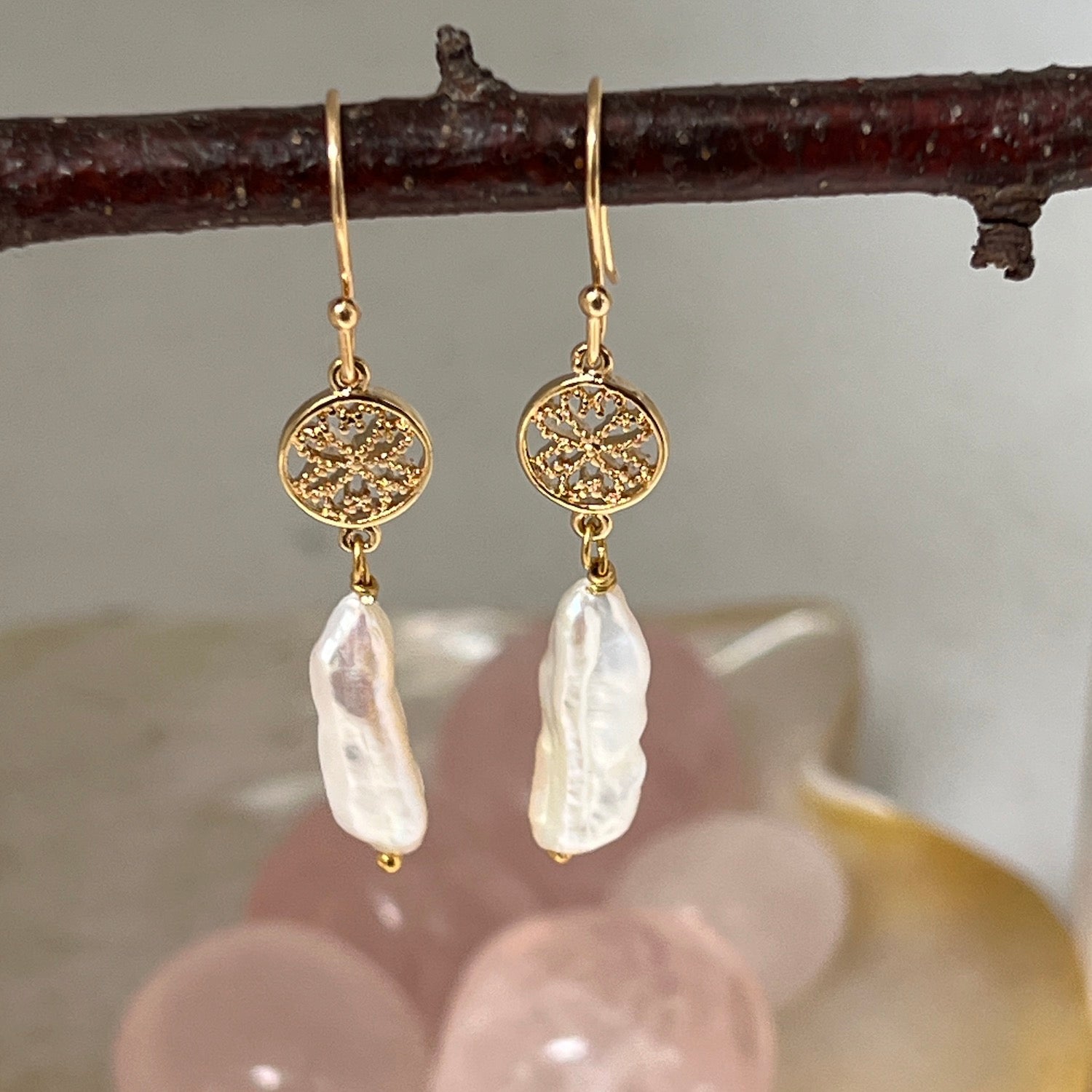 Close-up of a pair of gold-plated baroque pearl earrings, hanging from a twig with crystals in the background