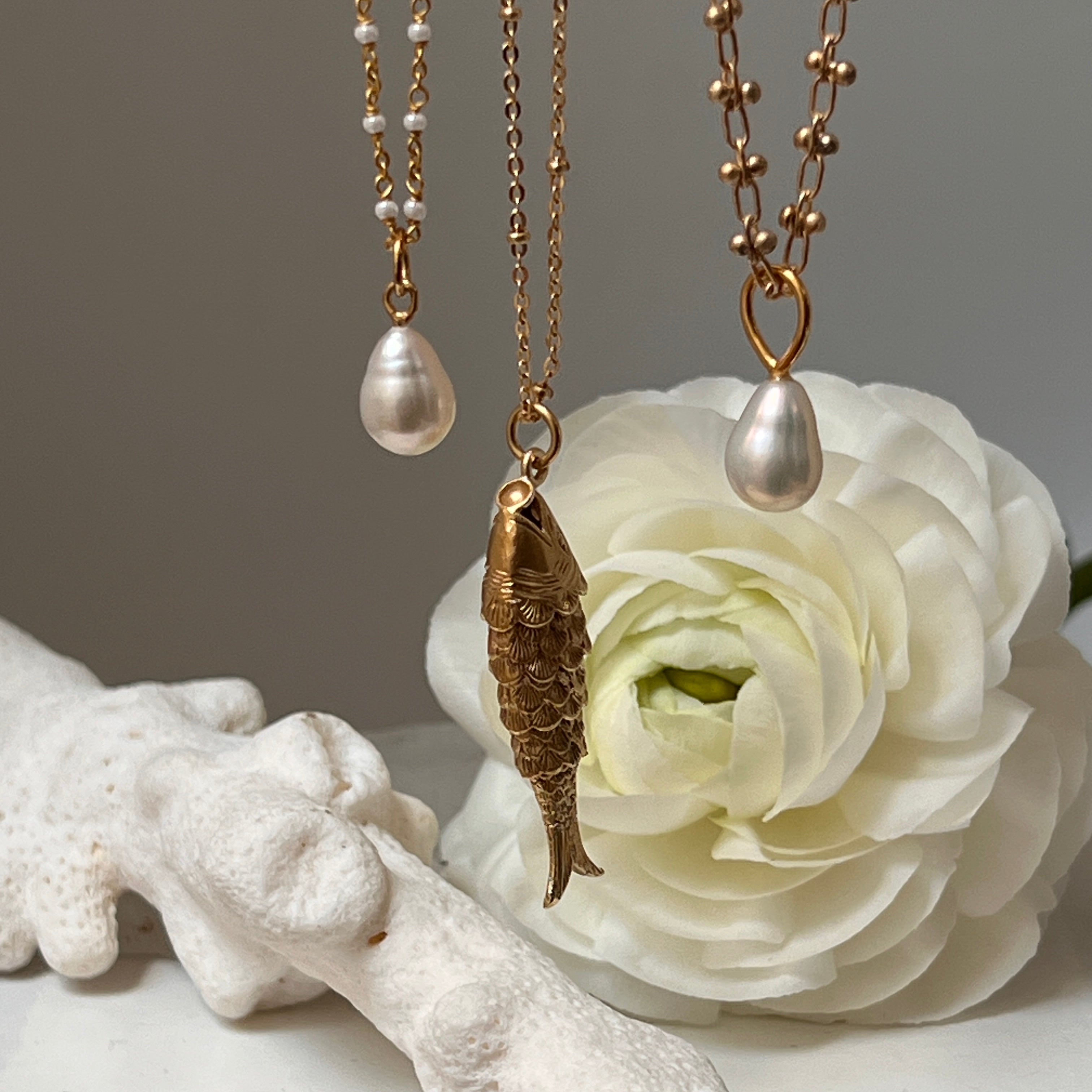 A gold-plated articulated fish charm on a gold satellite chain displayed with 2 other necklaces with a rose in the background