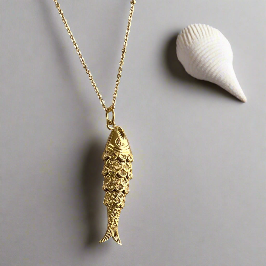 A gold-plated articulated fish charm on a gold satellite chain displayed with a seashell in the background