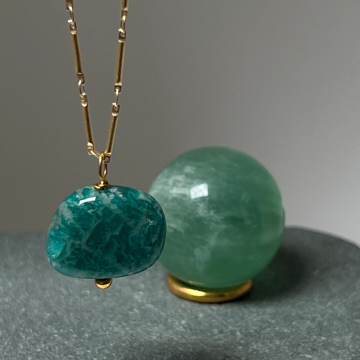 Green stone pendant on a gold chain with a matching green stone sphere on a gray surface from Mirabelle jewellery