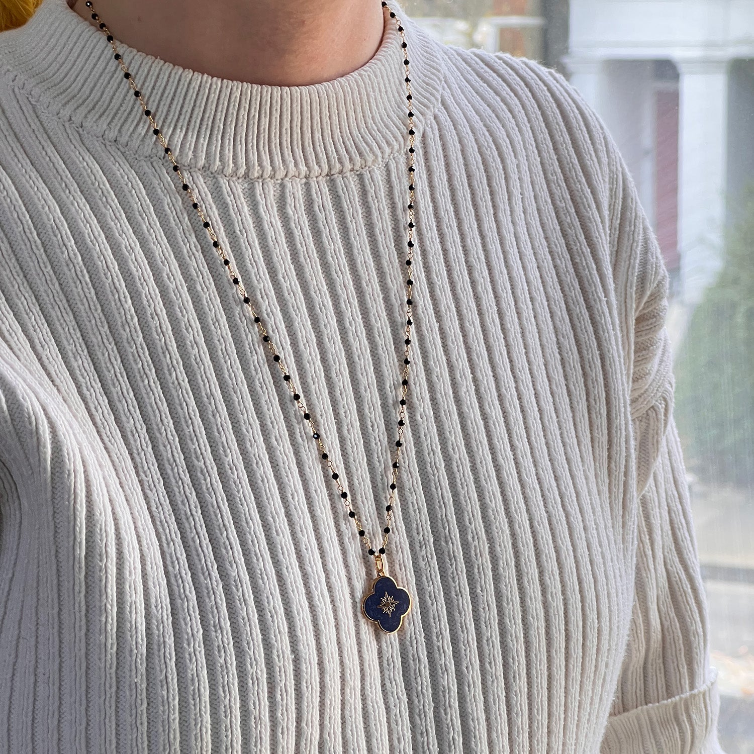 A royal blue enamel medal with a gold-plated brass cross symbol on the front, featuring a star design at the center, attached to a long black onyx rosy chain. Worn on a model wearing an ivory jumper.