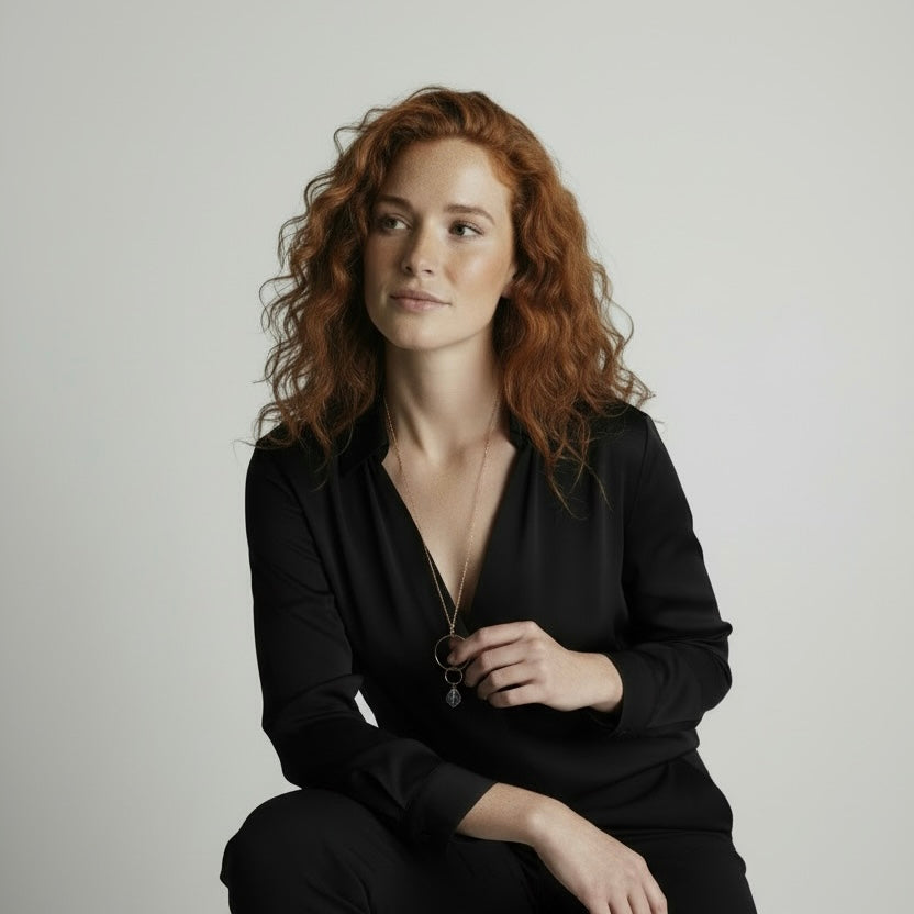 Woman with red hair wearing a black outfit against a plain background, wearing a gold necklace with a freeform rock crystal pendant.