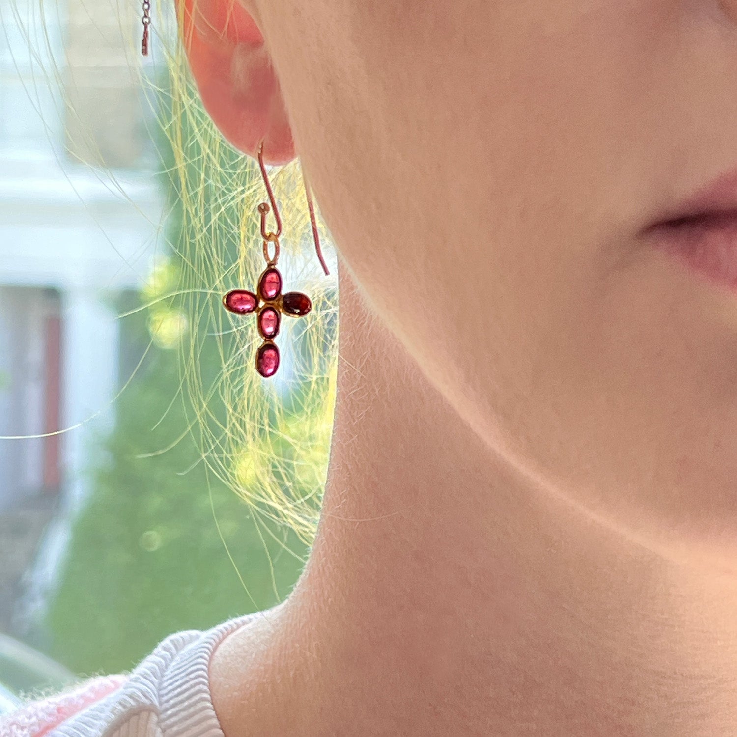 Close-up of a person wearing a garnet cross earring with a blurred outdoor background