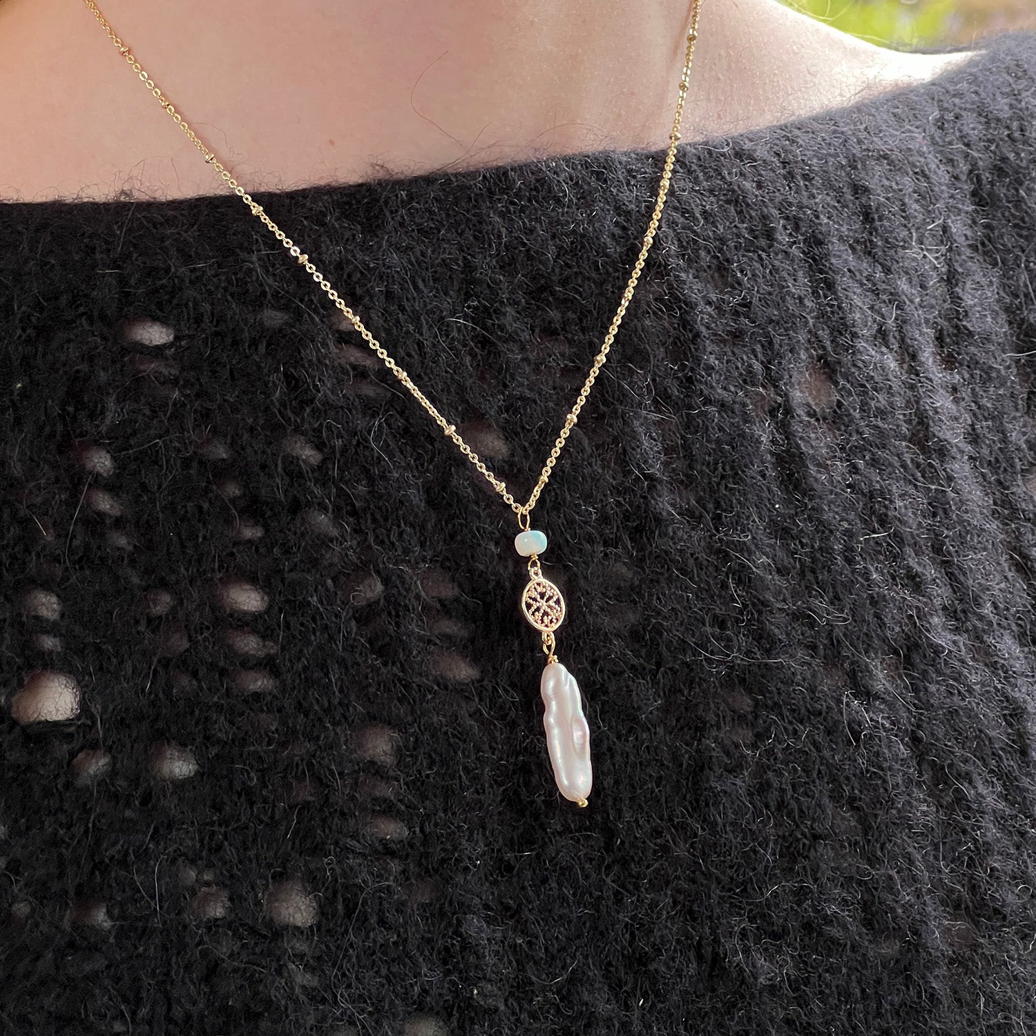 A gold-plated pendant featuring a baroque pearl and opal worn by someone wearing a black jumper