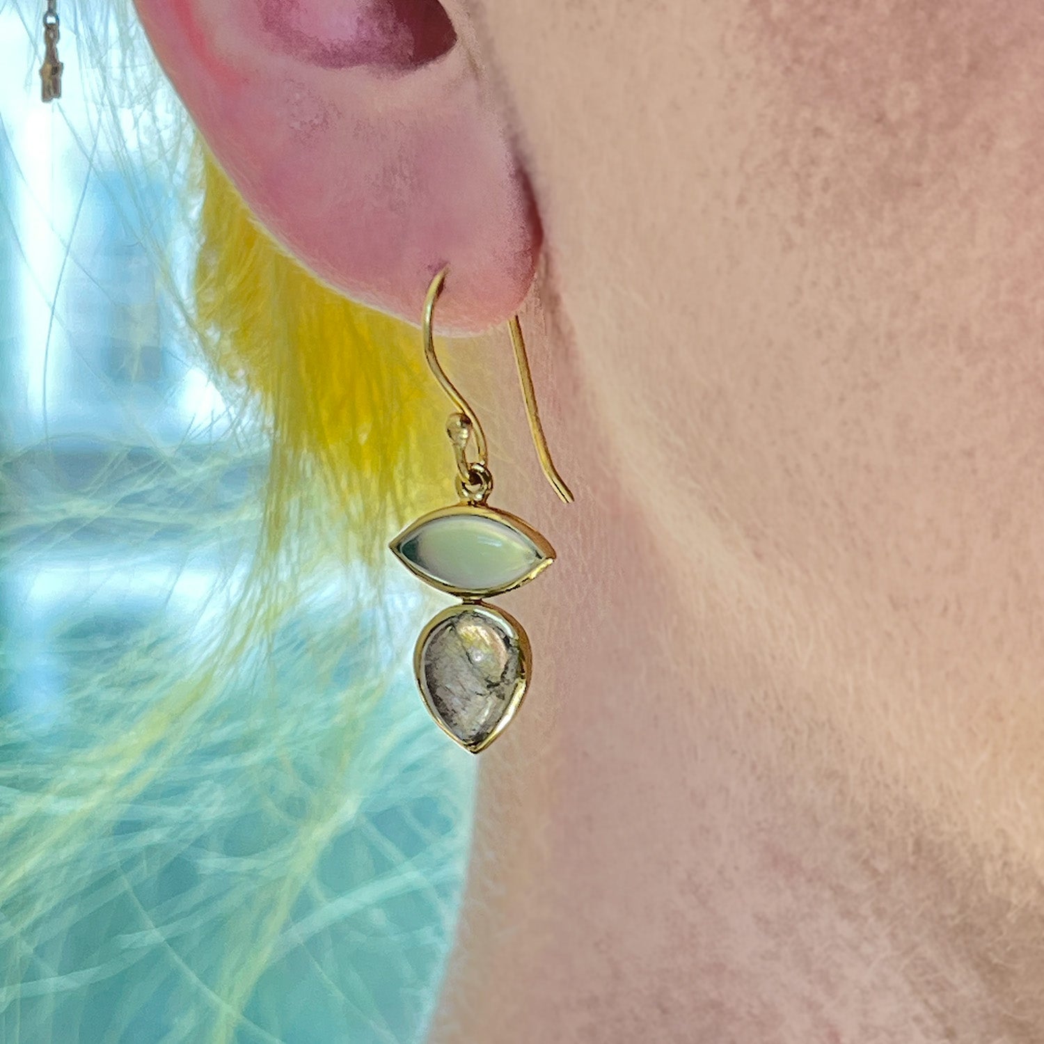 Gold-plated earrings with labradorite and aqua chalcedony gemstones worn by a person against a blurred natural background