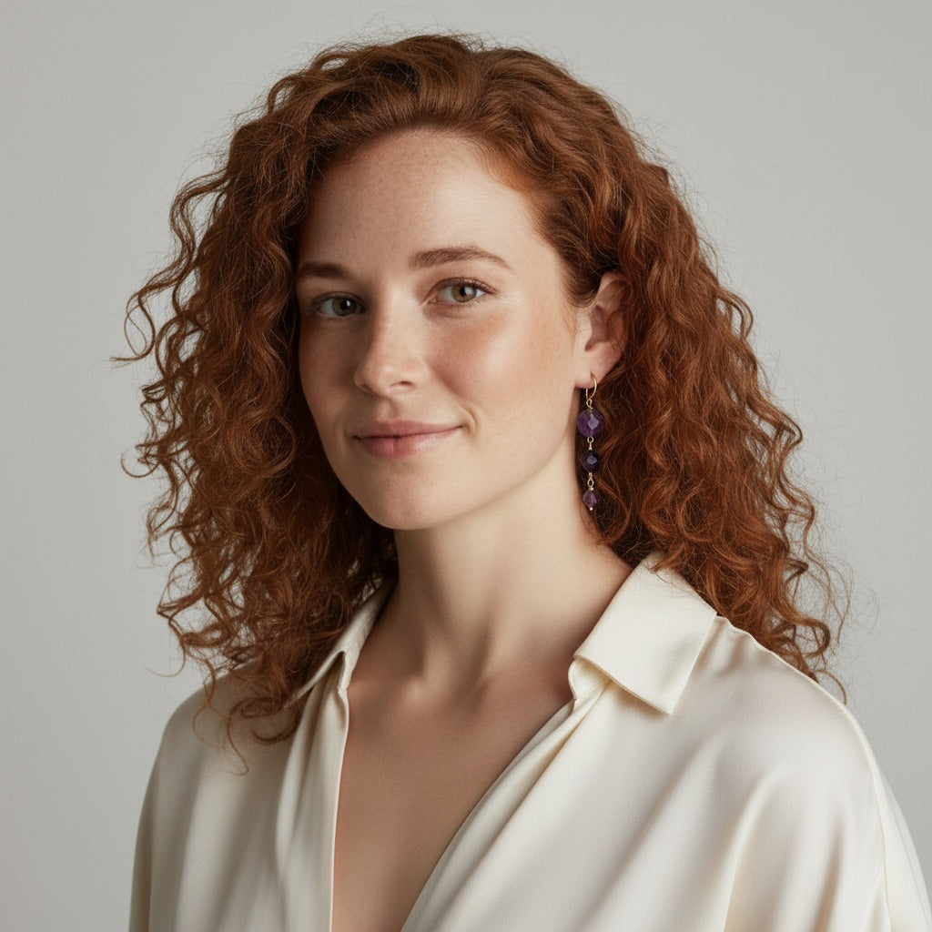 Woman with curly red hair wearing amethyst earrings with a white blouse against a neutral background