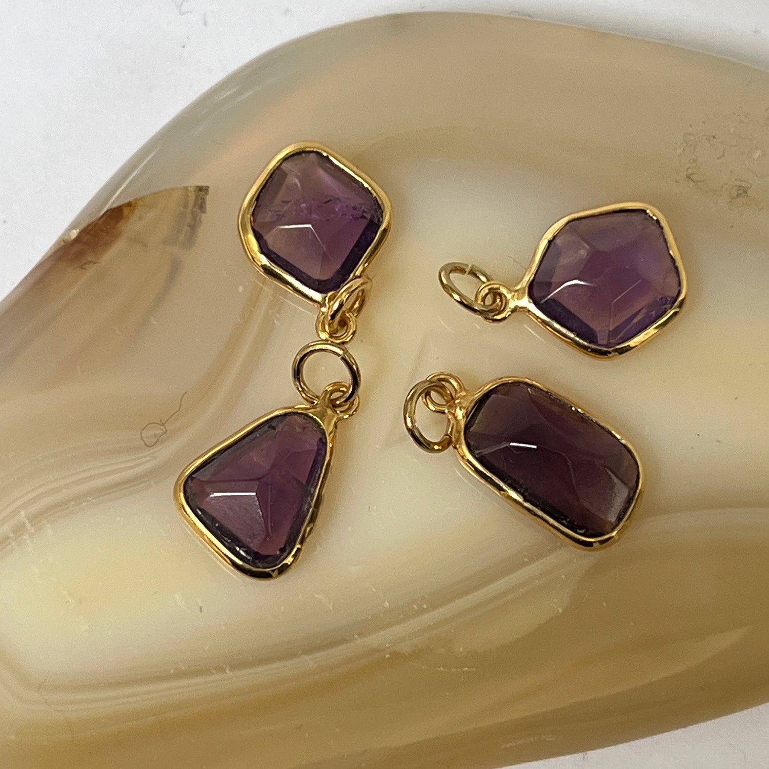 Four gold pendants with purple amethyst stones on a beige stone background