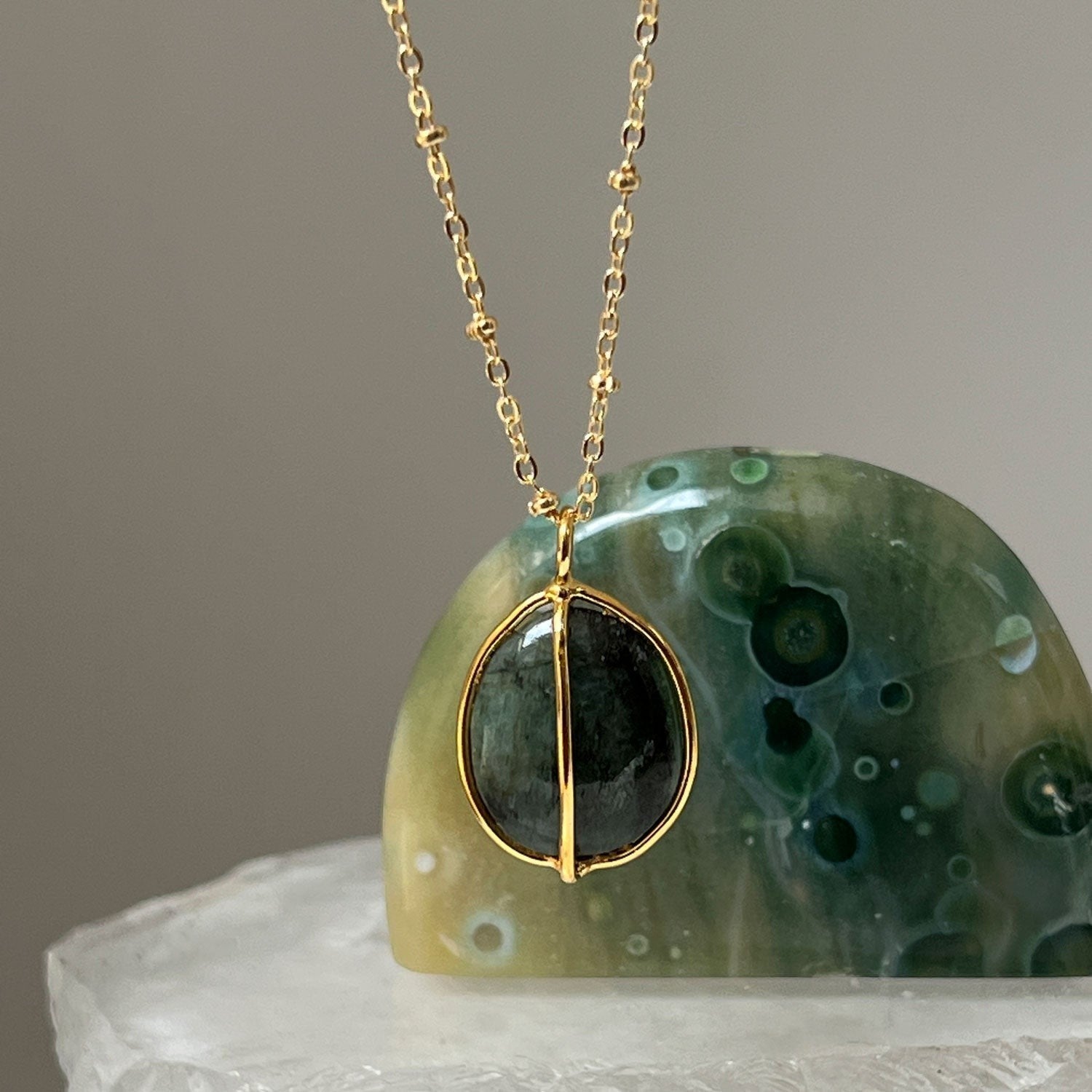 Mirabelle Jewellery freeform raw tumbled emerald pebble pendant on a gold satellite chain. The dark green stone has a natural, earthy texture.