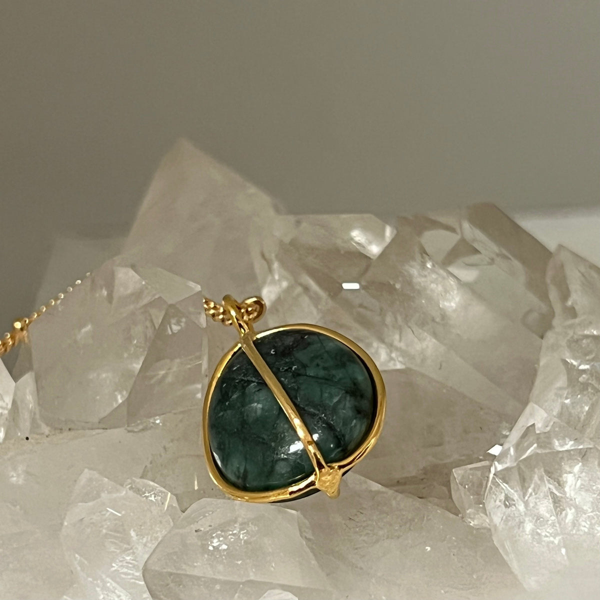 Mirabelle Jewellery freeform raw tumbled emerald pebble pendant on a gold satellite chain, set against a backdrop of clear crystals.