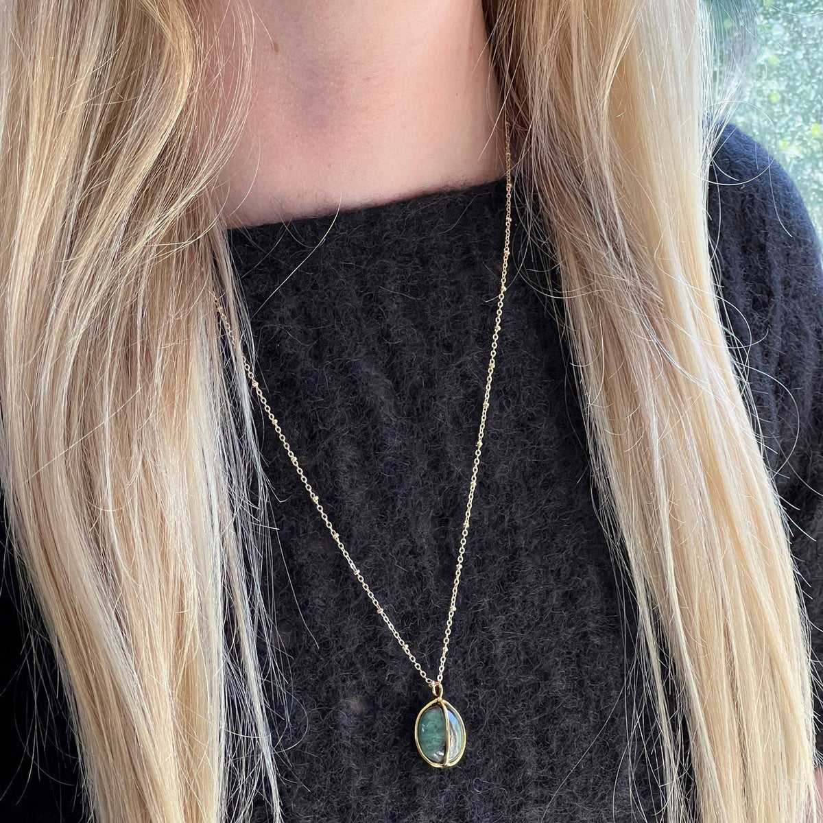 Mirabelle Jewellery gold satellite chain necklace with a freeform tumbled emerald pebble pendant. The pendant is oval-shaped with a raw, natural green emerald stone.