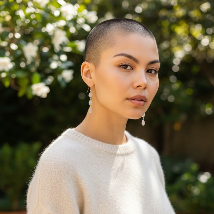 Woman with a shaved head wearing gold-plated rose quartz earrings with a white sweater outdoors