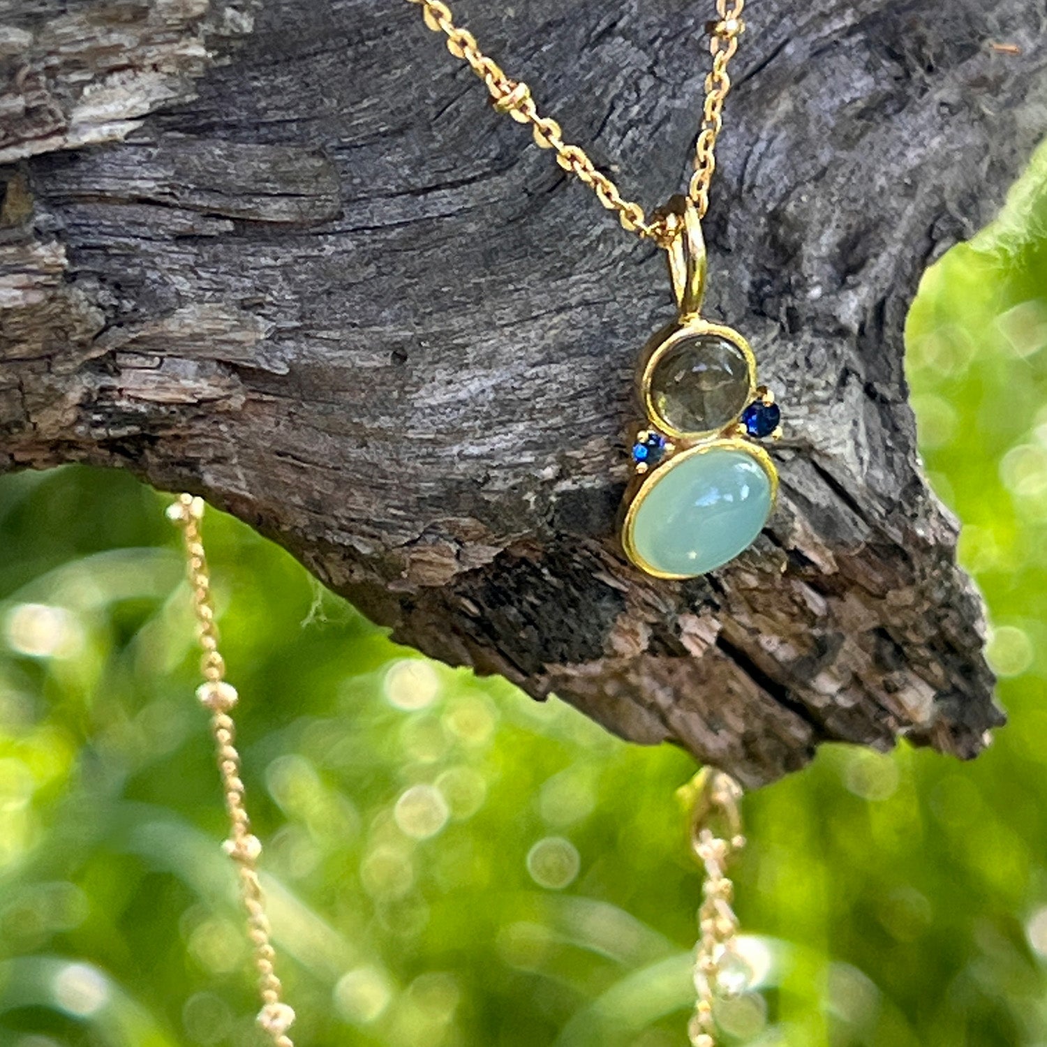 Pendant gold necklace with three stones: Aqua Chalcedony, Labradorite, and Blue Zircon on a wooden branch.