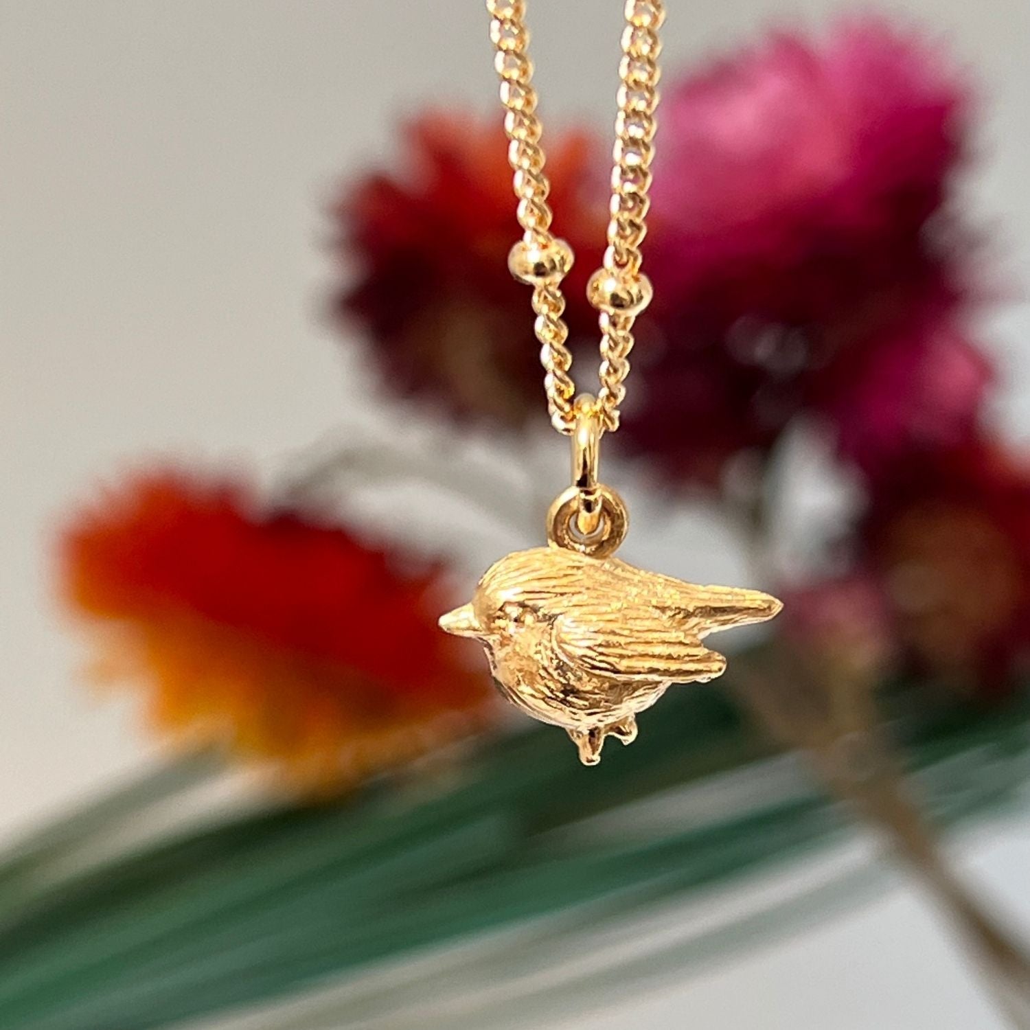 Elegant 22ct gold-plated necklace with a gold Biba chain and cute robin charm. Flowers are in the background