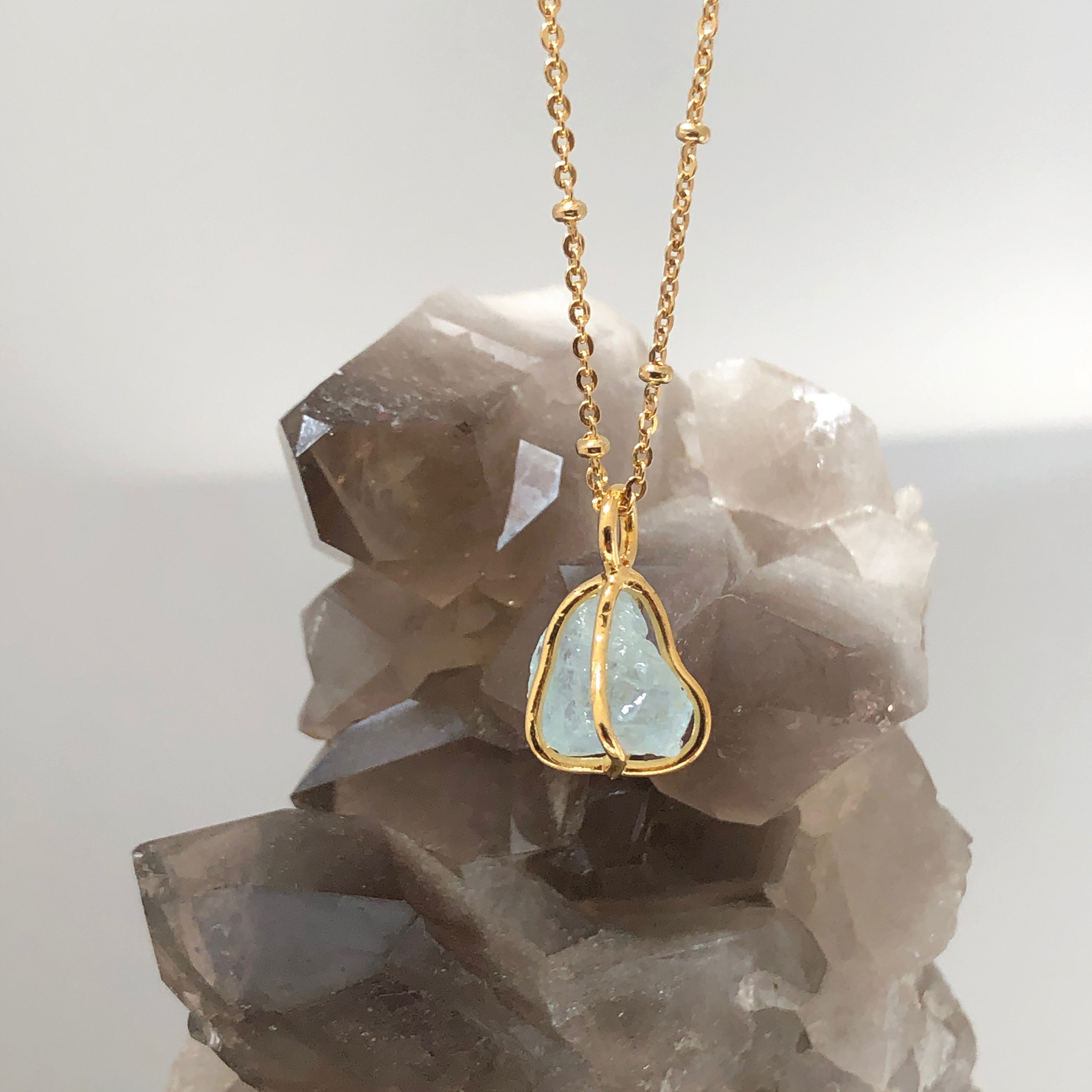 A gold-plated brass pendant with raw Aquamarine crystal encapsulated in a cage, attached to a short satellite chain necklace with crystals in the background