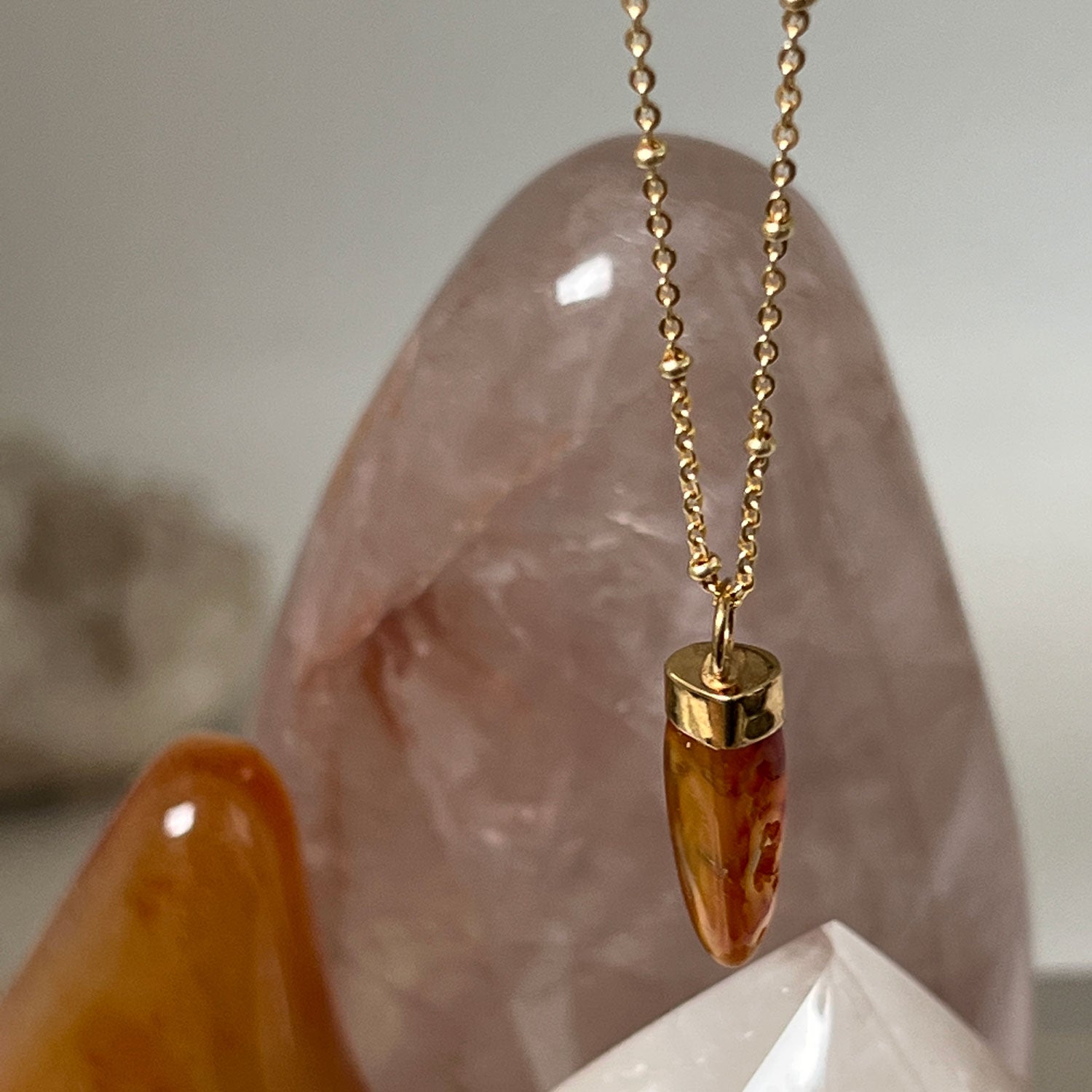 A close up of a gold-plated pendant with a carved orange Carnelian stone, with crystals in the background