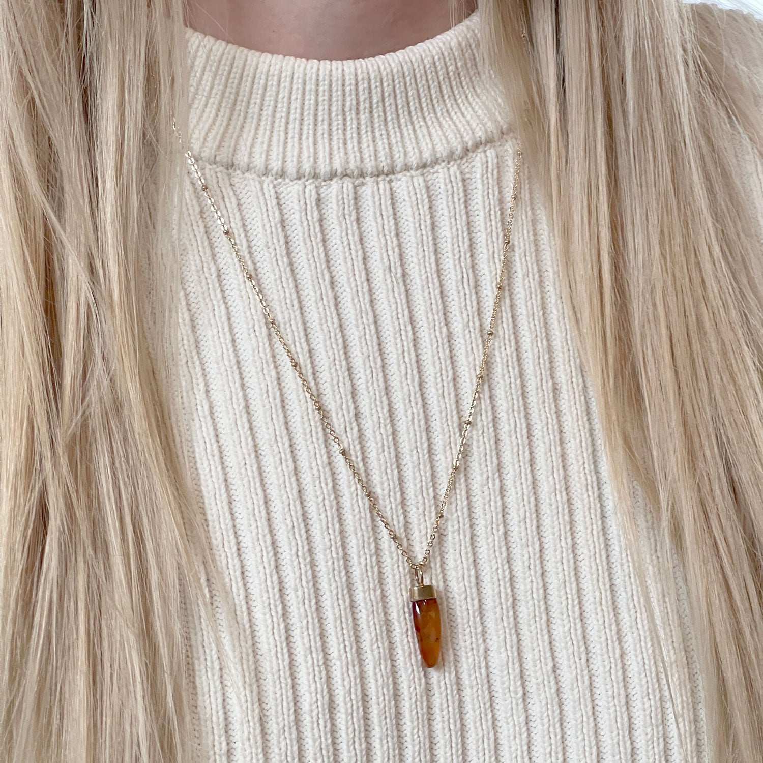 A gold-plated pendant with a carved orange Carnelian stone, attached to a long satellite chain. Worn around a model's neck on an ivory jumper.