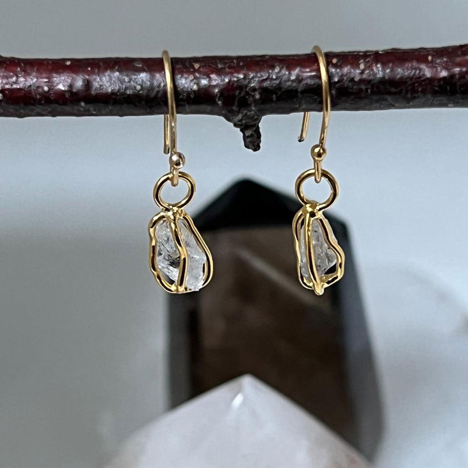 Raw Herkimer quartz diamond crystal earrings set in delicate gold-plated cages hanging from a twig.