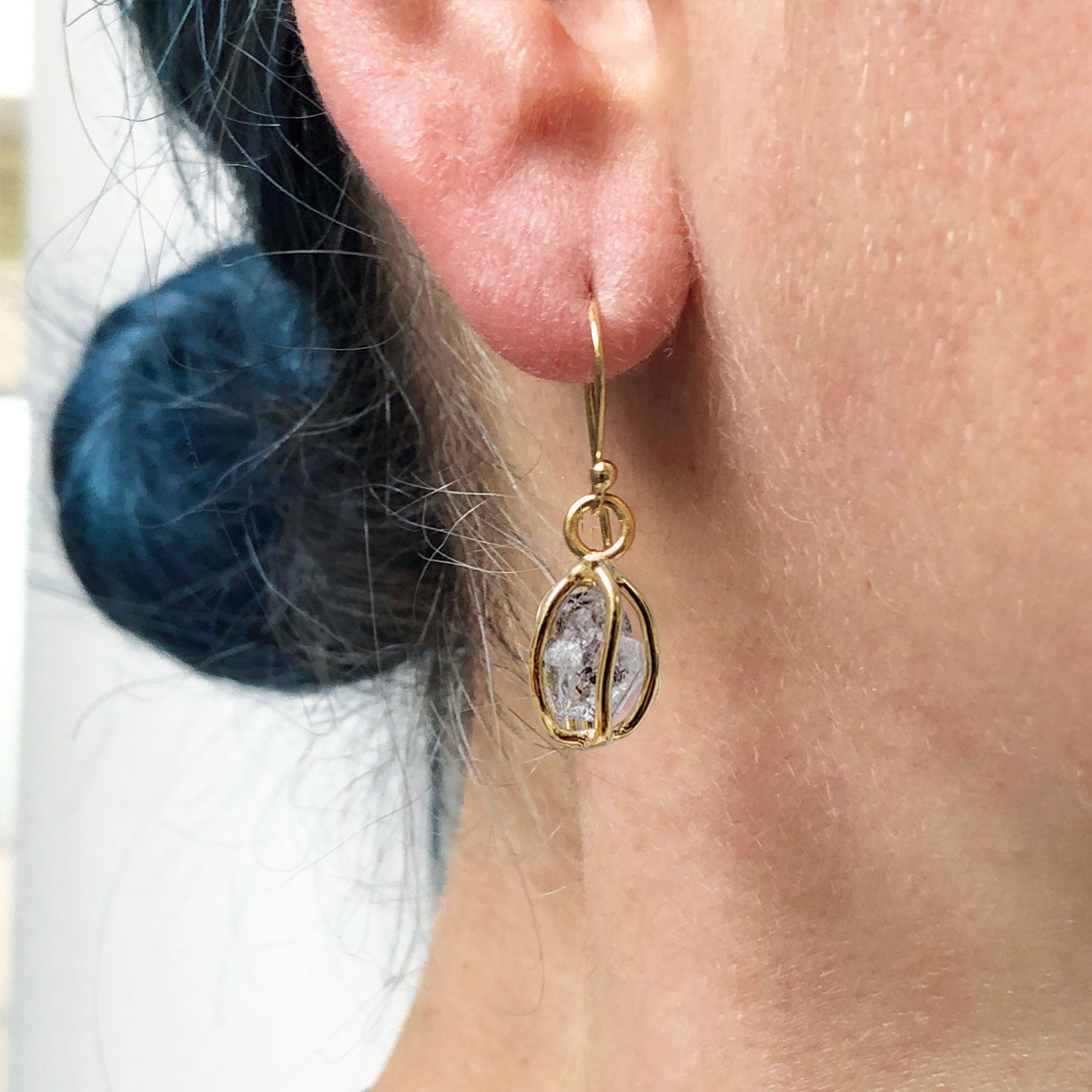 A close up of a model's ear wearing a Herkimer quartz diamond crystal earring set in delicate gold-plated cages.