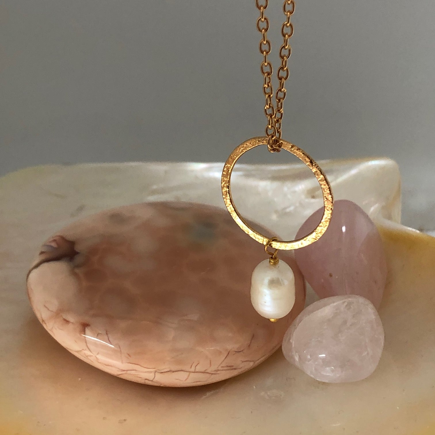 Close up of a freshwater pearl pendant, hanging from a gold-plated hammered ring and on a gold-plated chain. Coloured stones in the background.