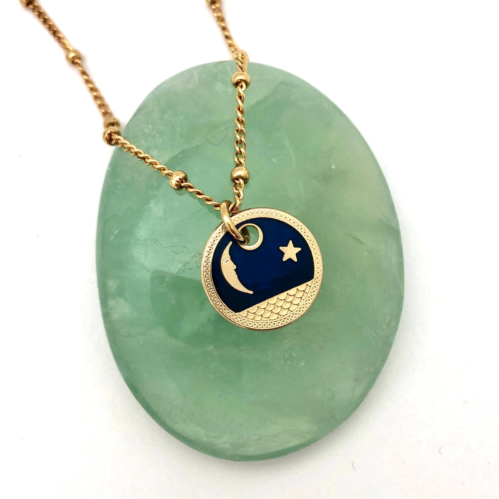 A royal blue enamel and gold-plated pendant, depicting the moon and a star, on a gold-plated chain. Draped over a green stone