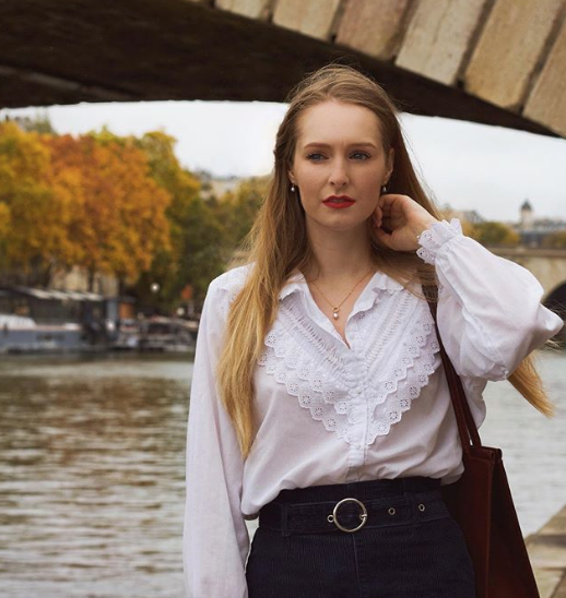 A model wearing a gold pink pearl necklace with a white blouse, standing next to a river