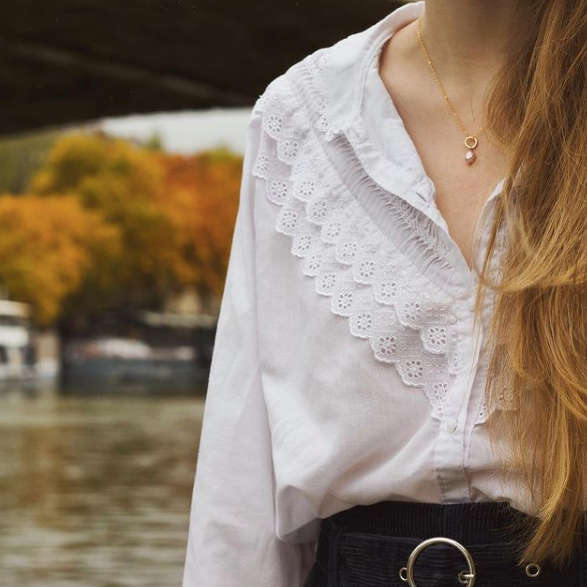 Close-up of a model wearing a gold pink pearl necklace with a white blouse, long hair, standing next to a river