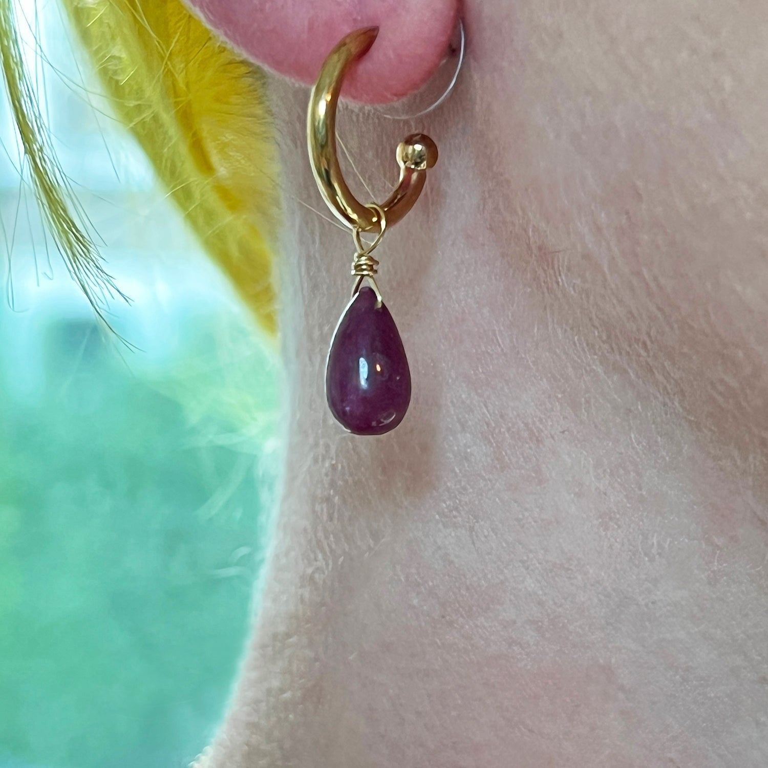 Gold earring with a ruby teardrop pendant worn on an ear against a blurred background