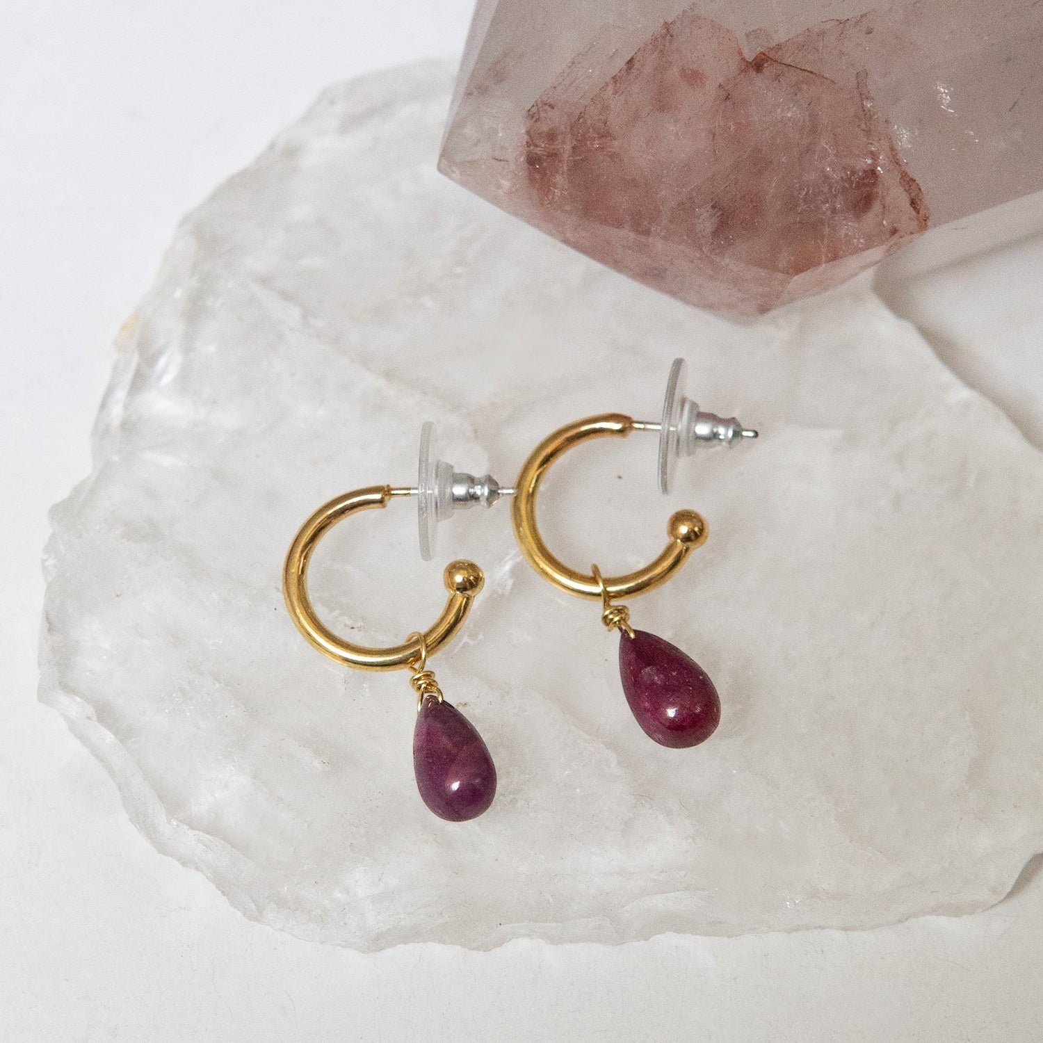Gold hoop earrings with ruby teardrop charms on a white shell and crystal background